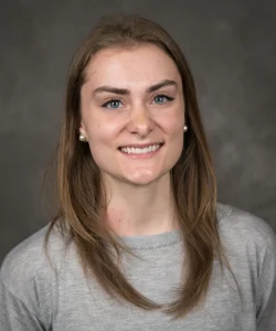 Aly smiles and has long light brown hair. Aly wears a grey blouse.