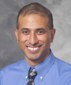 Manish Shah is smiling while wearing light blue. Manish has short, dark hair.