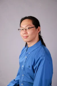 Yixuan Chen is smiling and wears a blue button-up shirt. He has long black hair and black glasses.