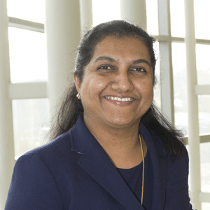 Dr. Mathew smiles for her photo. She has long, dark hair, and she wears a blue blazer.