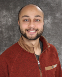 Nicholas Faulkner smiles brightly. He has very short dark hair and wears a red sweater.